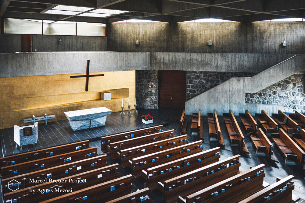 The interior of the Baldegg Kloster chapel. Wide angle view showing the wooden pews, the minimalist altar, and the dramatic board-marked concrete walls illuminated by natural light.