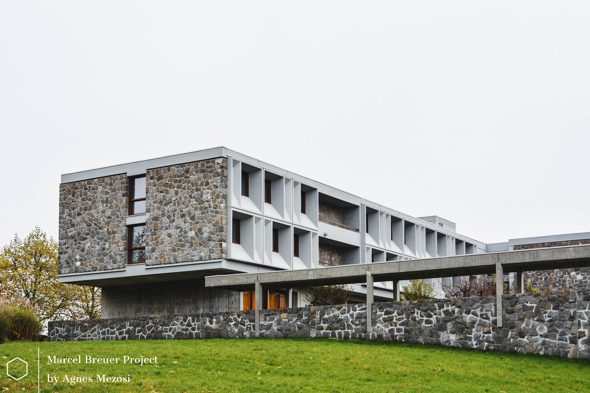Exterior view of the Baldegg Kloster in Switzerland by Marcel Breuer. A long, horizontal concrete and stone structure integrated into the green landscape under a bright sky.