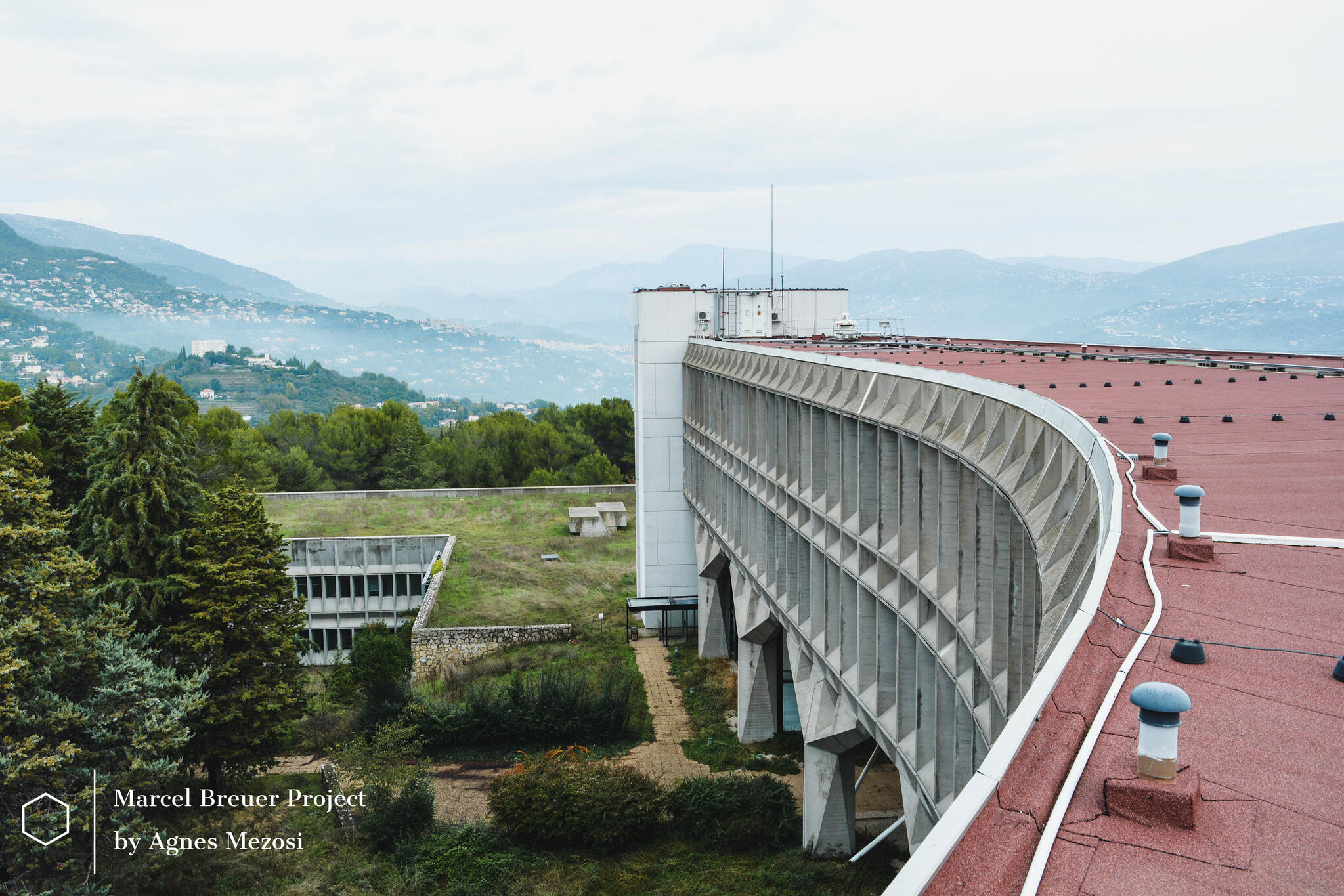 Aerial perspective of the curved concrete IBM Research Center building integrated into the French Riviera landscape with mountains in the background.