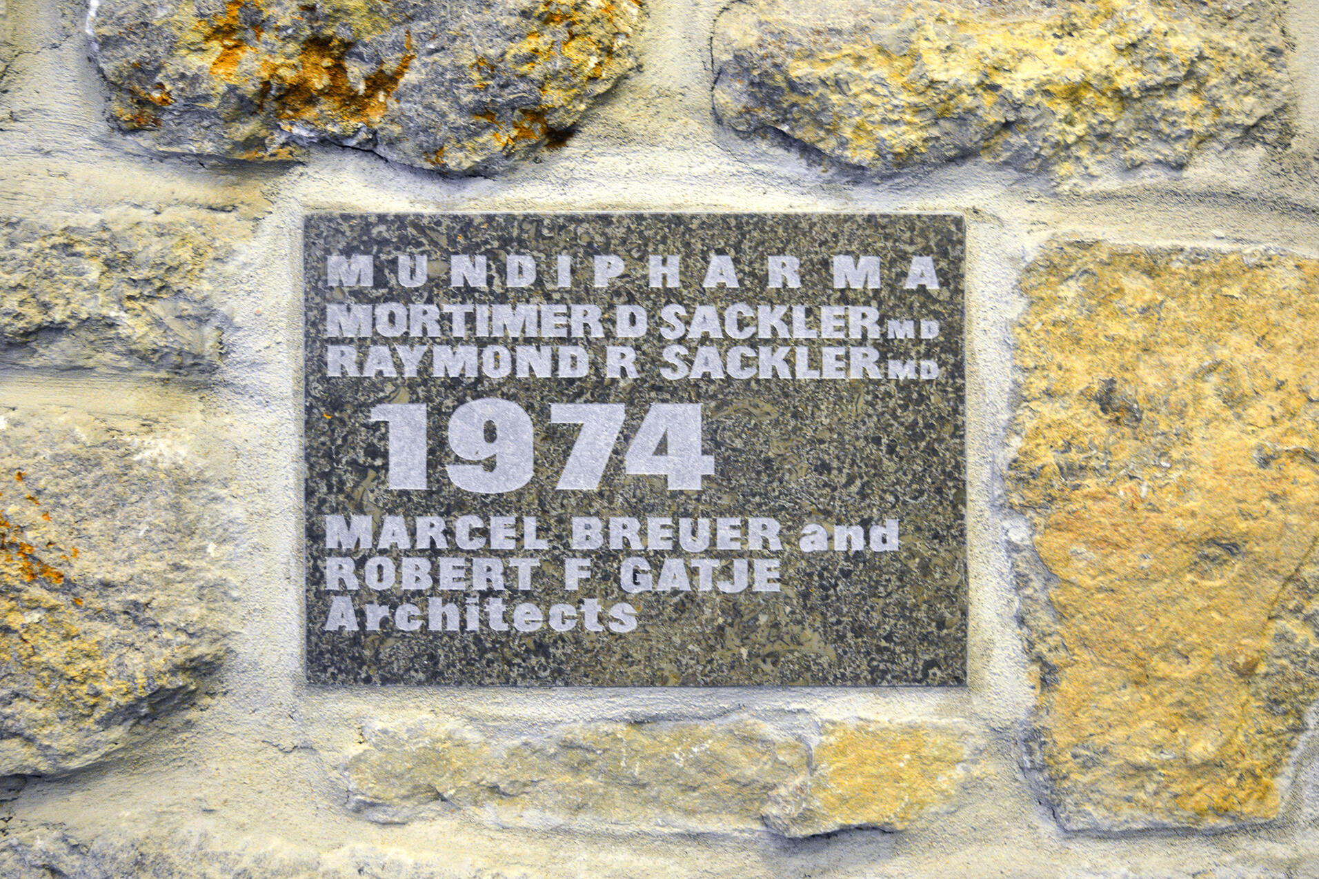 A granite commemorative plaque embedded in a rough stone wall, marking the 1974 completion of the Mundipharma building by Marcel Breuer and Robert F. Gatje.
