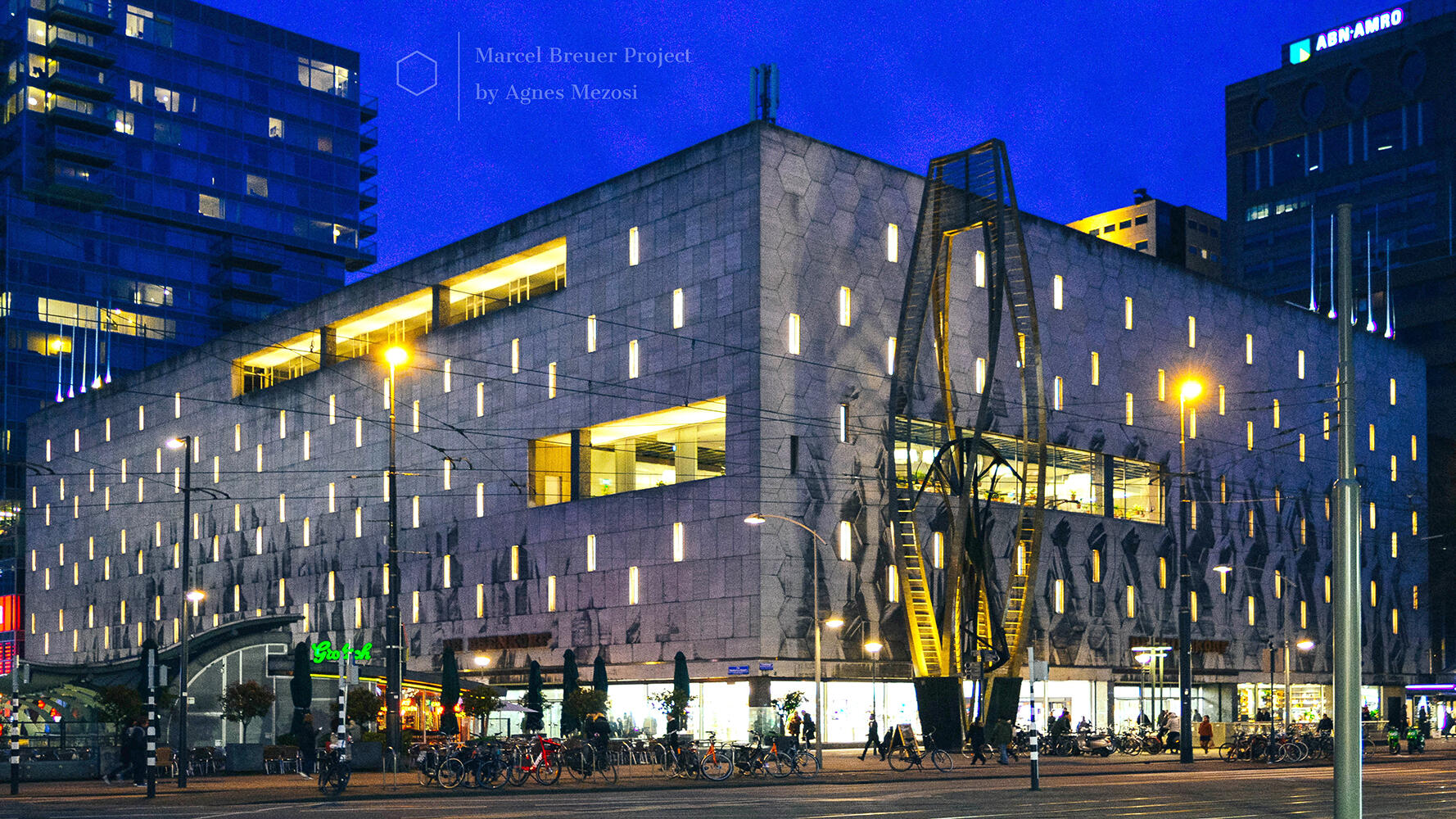Night photograph of the De Bijenkorf department store in Rotterdam, showing the massive stone facade dotted with small, glowing rectangular windows.