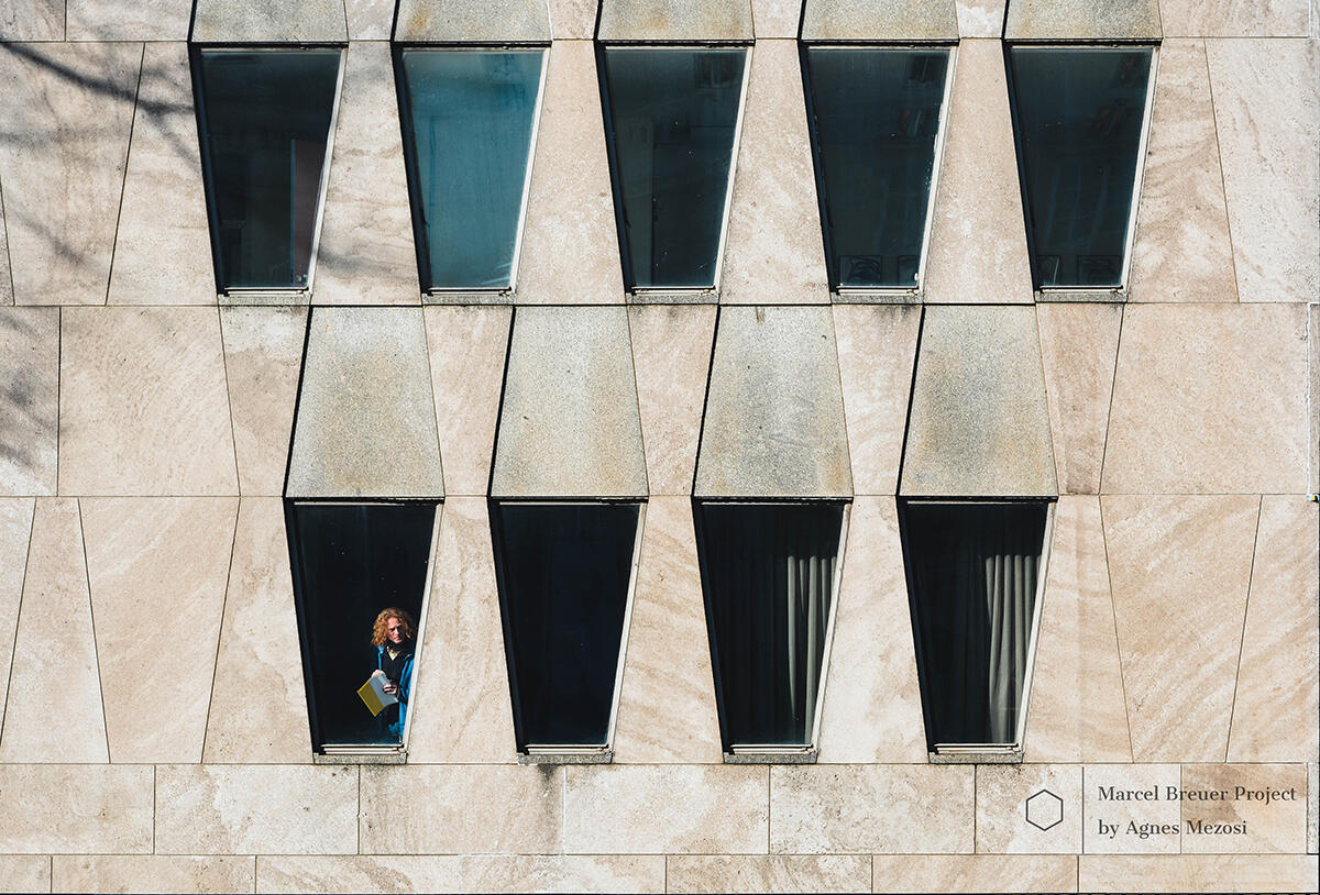 A color photograph of the embassy's facade where a person is visible behind one of the trapezoidal glass panes, surrounded by light beige stone panels.