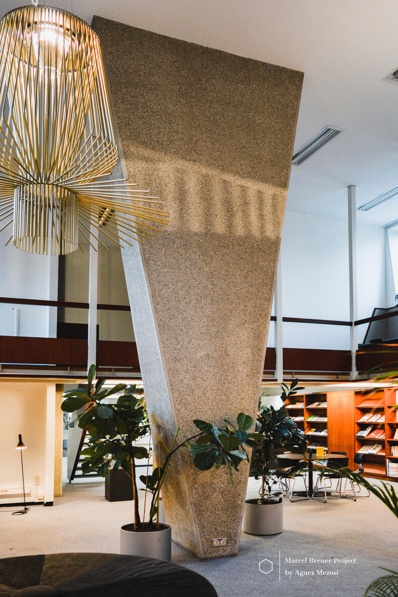 Interior color photograph of a dramatic concrete column with a twisted trapezoidal geometry that widens toward the ceiling, mirroring the angular patterns of the building's exterior facade.