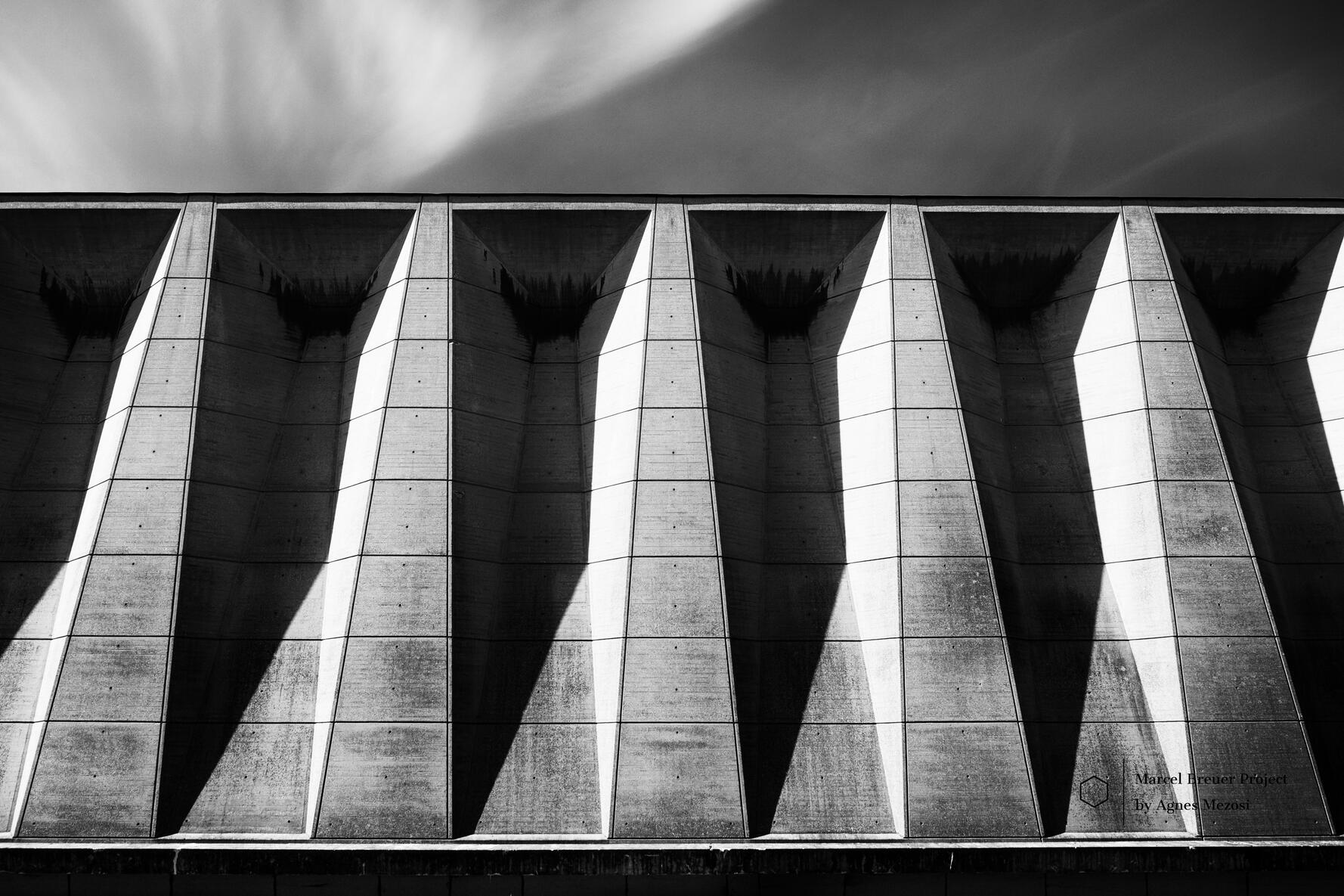 A wide, low-angle black and white photograph showing a repetitive row of massive concrete pillars under the building's overhang, creating a dramatic architectural colonnade.
