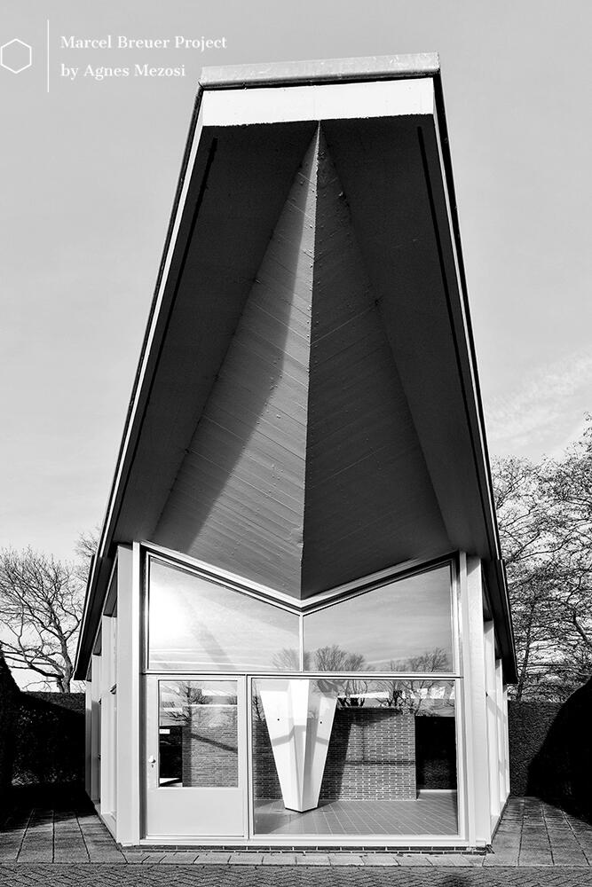 Black and white photograph of a small, a standalone, triangular modernist gatehouse with a sharp overhanging roof, part of the Van Leer site.