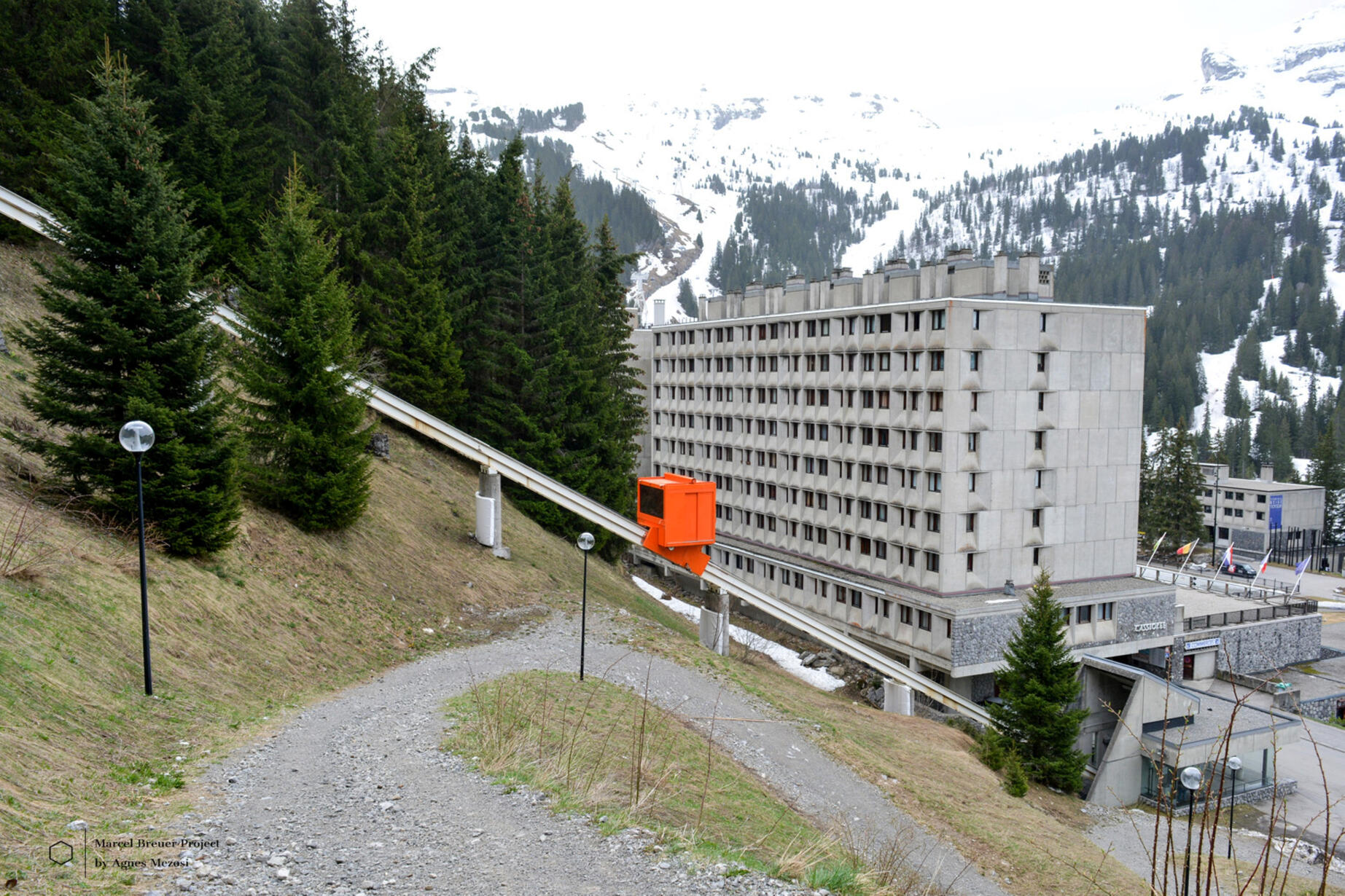 Wide shot of a large brutalist apartment block in Flaine with a bright orange cable car passing by on a forest slope.