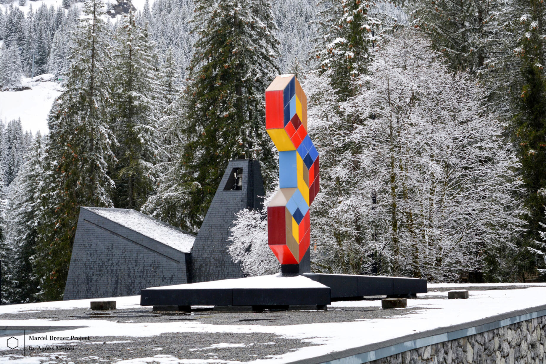 A vibrant, multi-colored geometric sculpture by Victor Vasarely in Flaine, with Marcel Breuer's dark concrete chapel in the background.