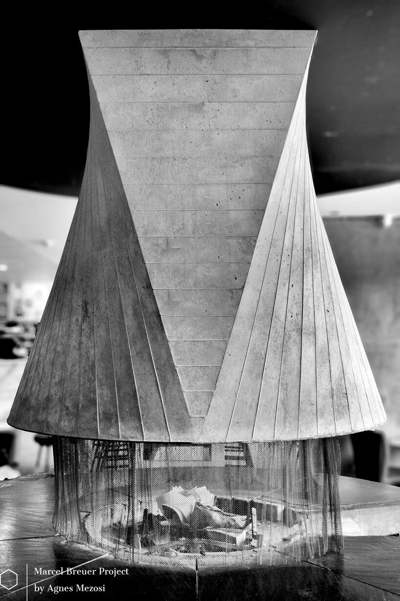 Interior black and white photograph of a large, sculptural concrete fireplace suspended from the ceiling.