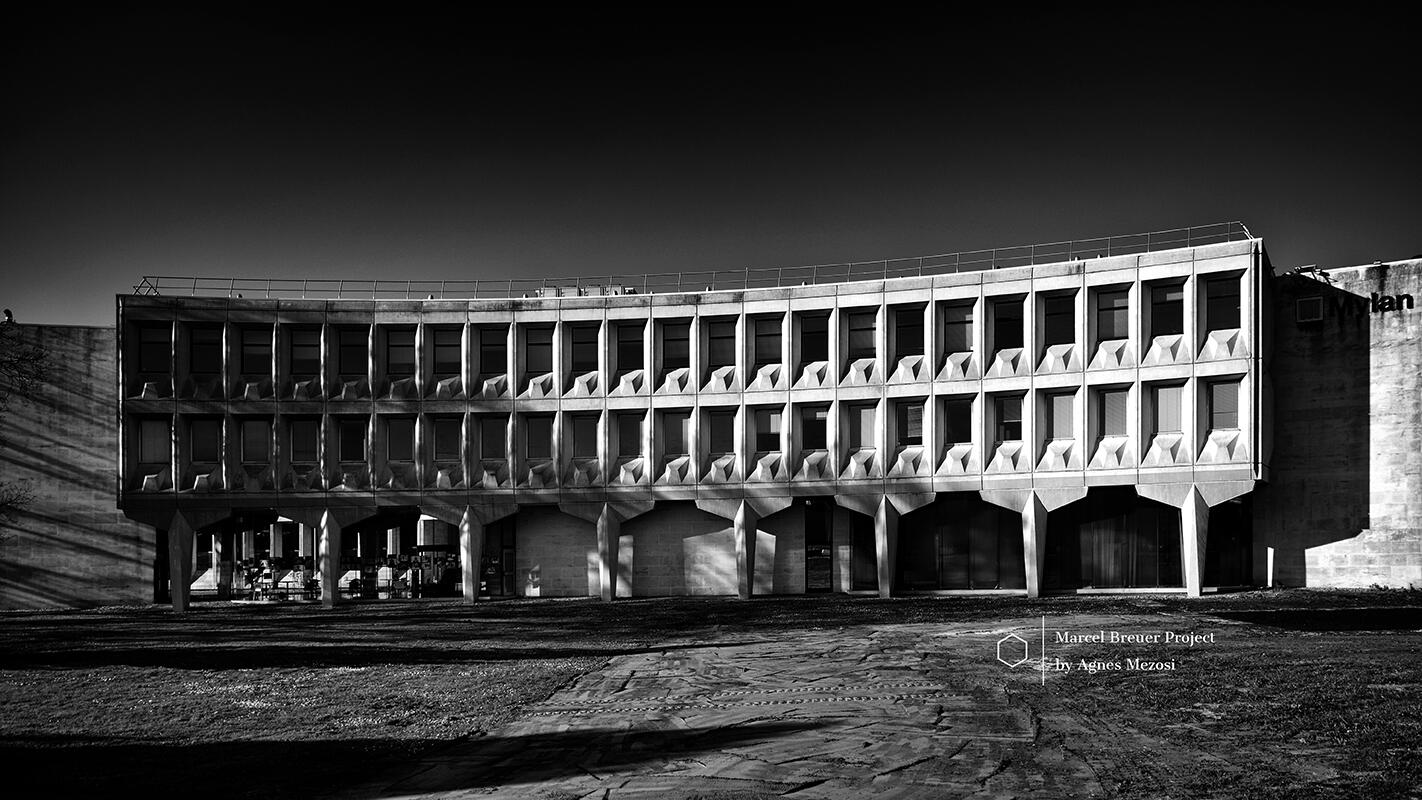 A black and white photograph of the Sarget-Ambrine building's main entrance facade, featuring a dramatic curved concrete structure supported by pillars.