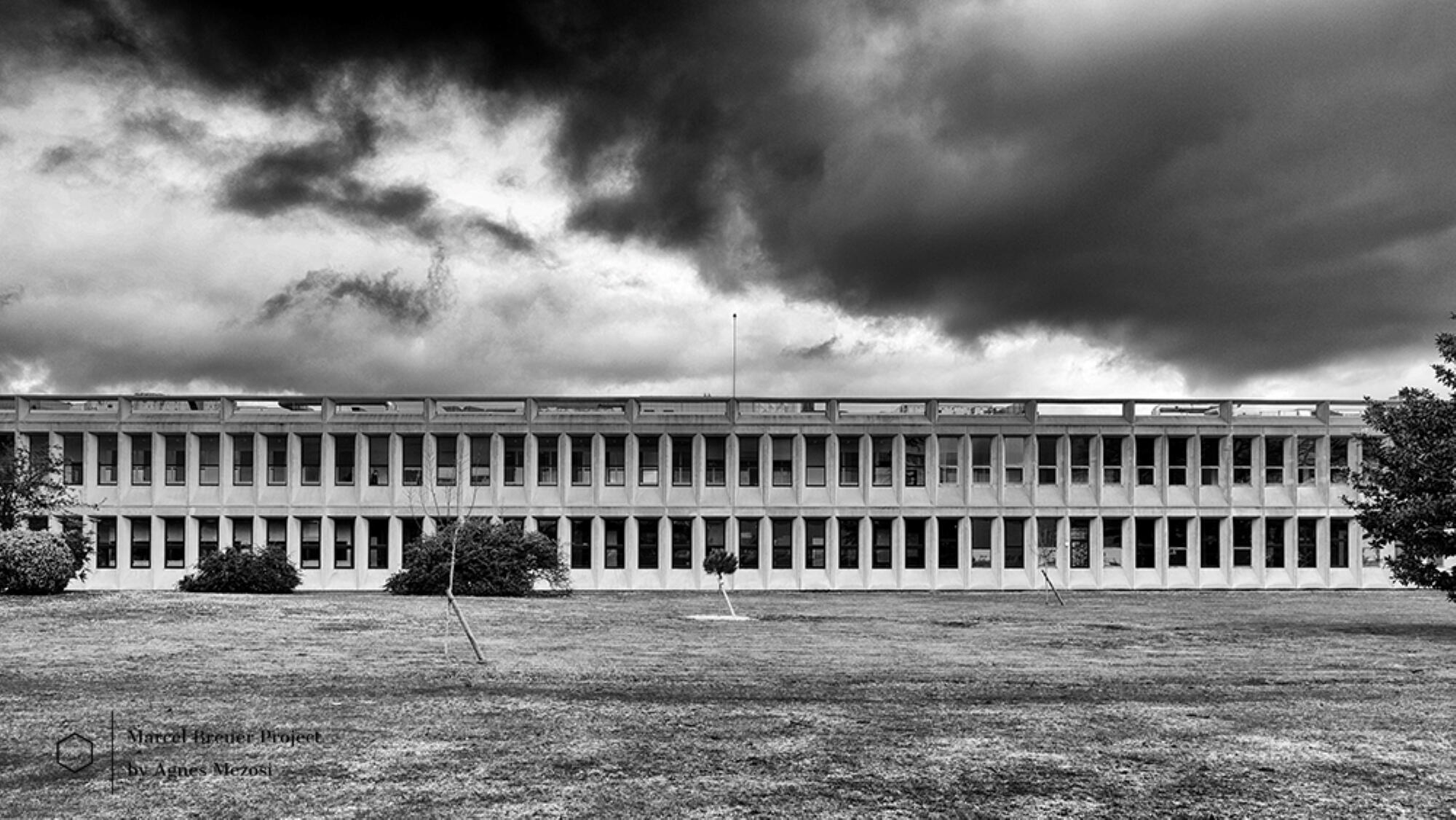 Wide black and white shot of the long, two-story Sarget-Ambrine concrete building under heavy, dark storm clouds.