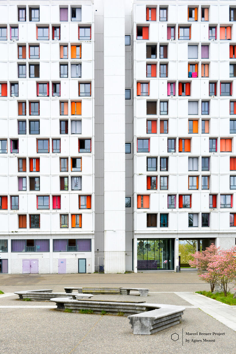A vertical shot of a tall residential building section with colorful accents, overlooking a paved courtyard with concrete benches.