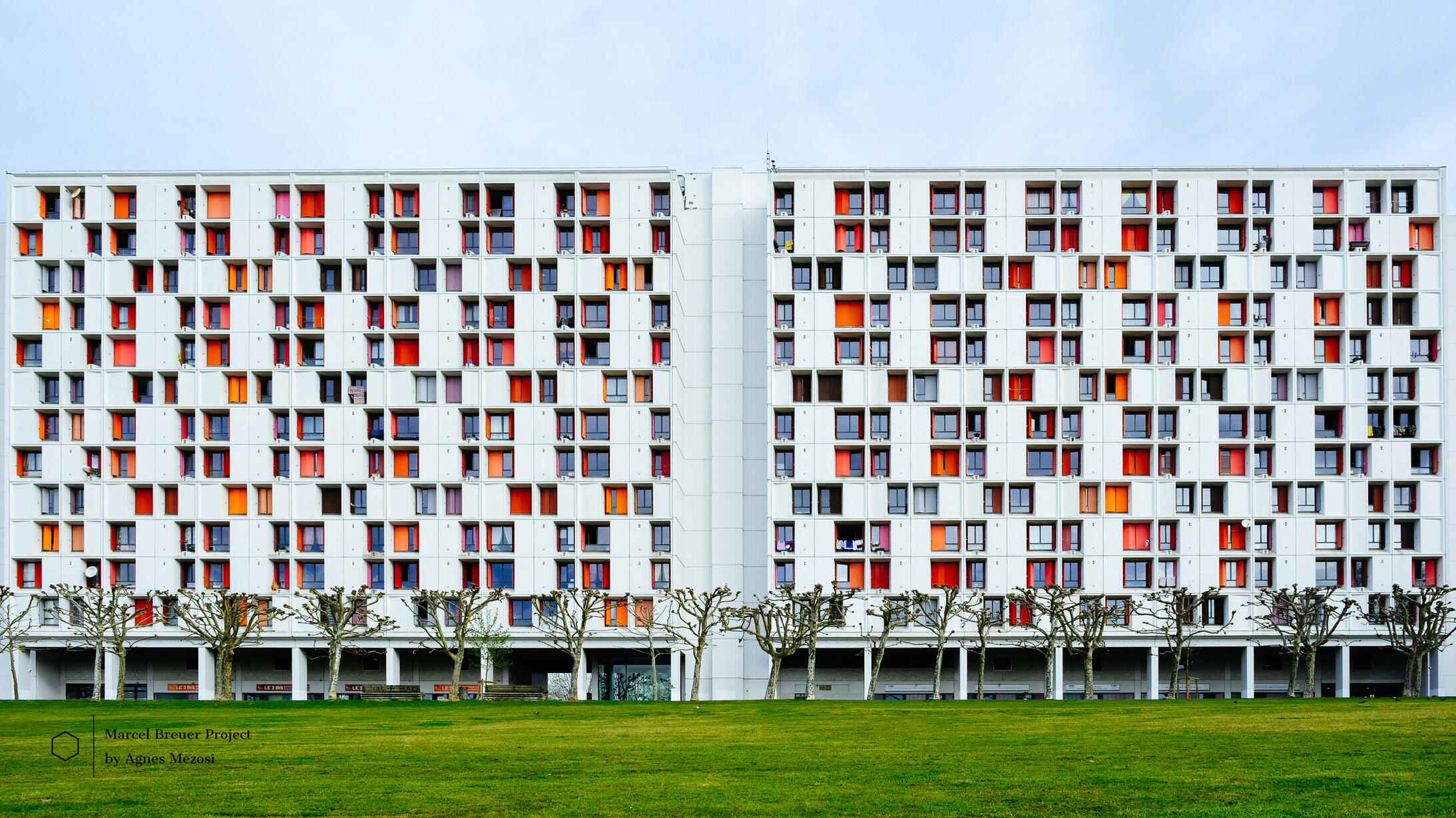 A wide-angle color photograph of a long, multi-story apartment block in Bayonne, featuring a repetitive grid of windows with orange, red, and blue panels.