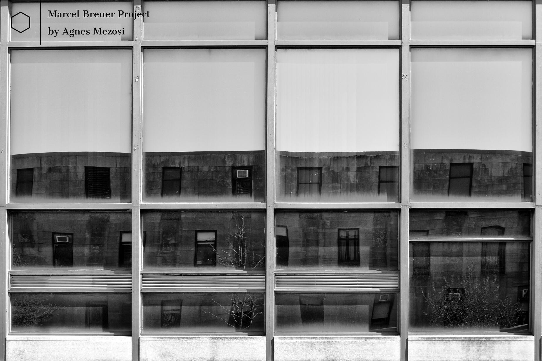 A black and white photograph showing the Shuster Hall's geometric concrete facade reflected perfectly in the large glass panes of the Fine Art Building.